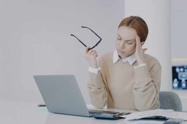 办公室桌子旁疲惫的年轻女子摘下了眼镜。员工在工作场所头痛。图片下载