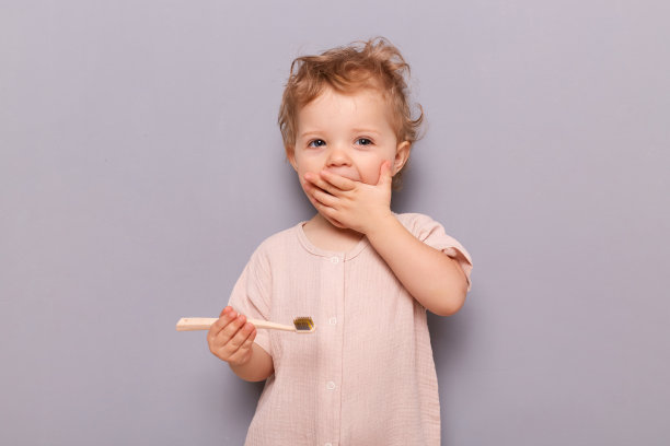 儿童牙齿护理。兴奋惊奇的小婴儿刷牙孤立在灰色的背景下，手里拿着牙刷，穿着休闲的童装站着。图片下载