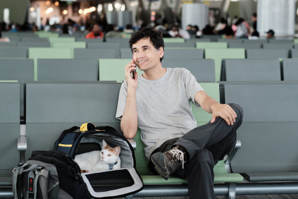 一名拉丁裔成年男子微笑着用手机说话，他坐在机场候机室他的猫旁边图片下载