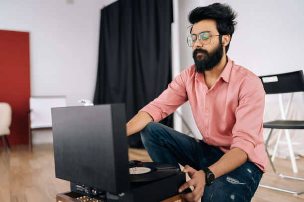 帅气的印度潮人戴着眼镜坐在地板上用旧唱机和黑胶唱片听音乐。音乐爱好者听古典音乐图片下载