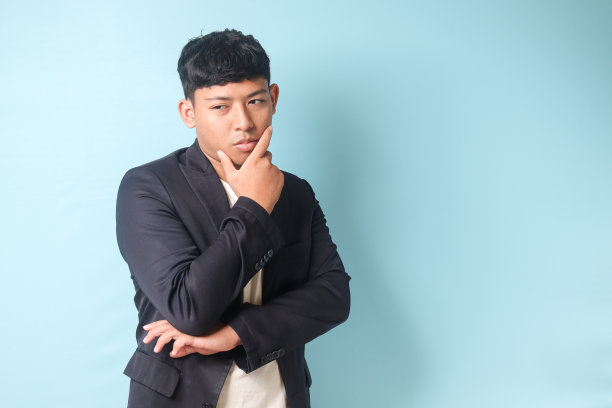 穿着休闲装的年轻亚洲商人的肖像，双手交叉在一起思考着什么。蓝色背景上的孤立图像图片下载
