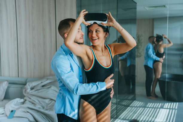 穿着泳装的开朗的西班牙年轻女子去掉了虚拟现实，在卧室和浴室里看着丈夫。大胡子在家里拥抱妻子。女人心存感激，收到礼物。图片下载