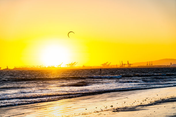 这张令人惊叹的照片捕捉到了日落时的海滩，金色的光芒照亮了海水和沙滩。在背景中，风筝冲浪者的剪影映衬着橙色和粉色的天空，创造了一个充满活力的海边冒险场景。图片下载