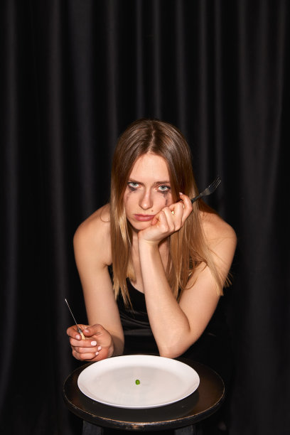 一个年轻苗条的女孩，时尚模特，坐在桌子旁，开始吃几乎空盘子的黑色背景图片下载