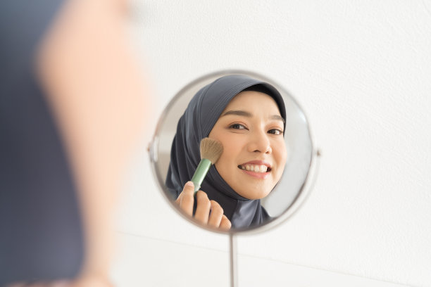 亚洲年轻的穆斯林妇女边照镜子边用刷子化妆。亚洲穆斯林女性戴着头巾，在脸颊上涂着化妆品睫毛膏。穆斯林妇女用腮红在颧骨上化妆图片下载