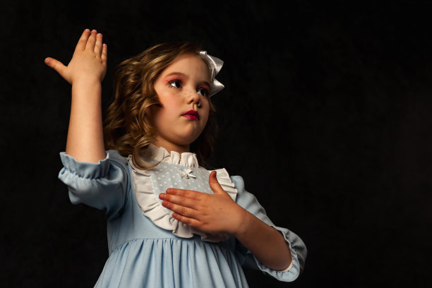 迷人的小娃娃封面女郎在黑色的背景下手势，看向别处。可爱的孩子穿着蓝色的衣服，头上戴着白色的蝴蝶结，与光和影隔绝。时尚形象理念。复制文本空间图片下载