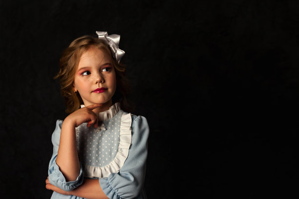封面小女孩的肖像，娃娃的形象在黑色背景，沉思地看着别处。工作室拍摄的可爱的孩子穿着蓝色的裙子与光和阴影隔离。时尚美容理念。复制文本空间图片下载