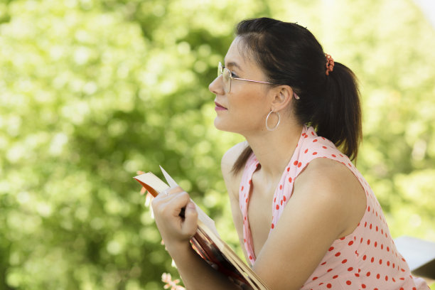 美丽的年轻女子戴着眼镜在房子的阳台上看书，春夏以自然为背景。一个正在看书的女人从阳台上若有所思地看着街道。本空间图片下载