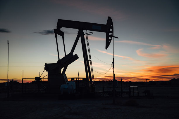 这张照片捕捉了一个油泵的轮廓，映衬在色彩斑斓的日落背景下。天空的暖色调与工业机械形成了美丽的对比，形成了独特而引人注目的形象。图片下载