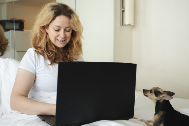 远程工作，女人在家工作，在笔记本电脑上打字，可爱的狗睡在床上，自由职业者图片下载