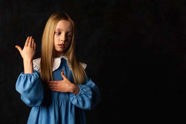 封面小娃娃女孩的肖像，穿着蓝色的裙子，在黑色的背景上做手势，看着别处。图片中9-10岁的可爱孩子被隔离在黑暗中。时尚化妆舞会概念。复制广告文本空间图片下载