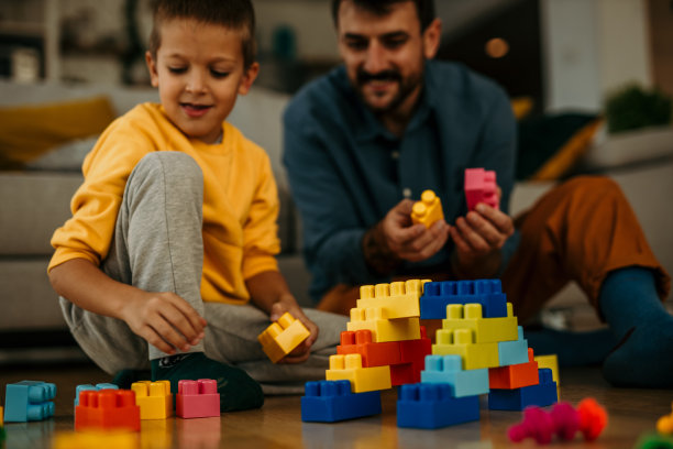 一位父亲和一个小男孩坐在幼儿园的地板上玩五颜六色的塑料积木。图片下载