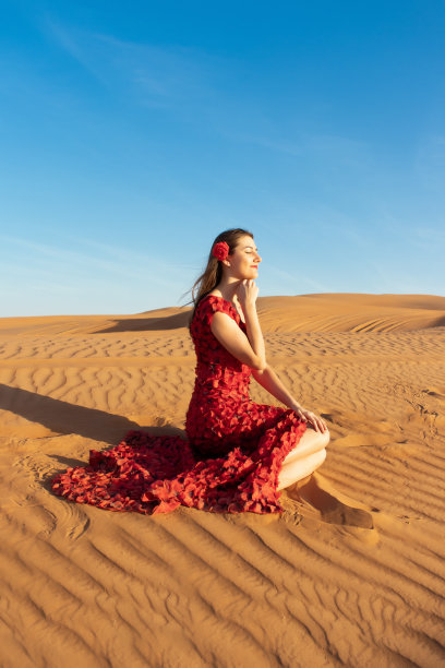 身穿红色长裙的年轻漂亮女人，沙漠中散落着红色玫瑰花瓣。沙漠玫瑰概念图片下载
