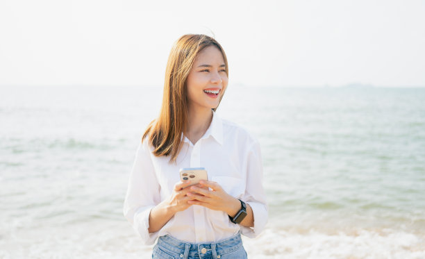 美丽的亚洲女人站在海滩上，手里拿着智能手机，在互联网上使用社交媒体。夏日生活理念。图片下载