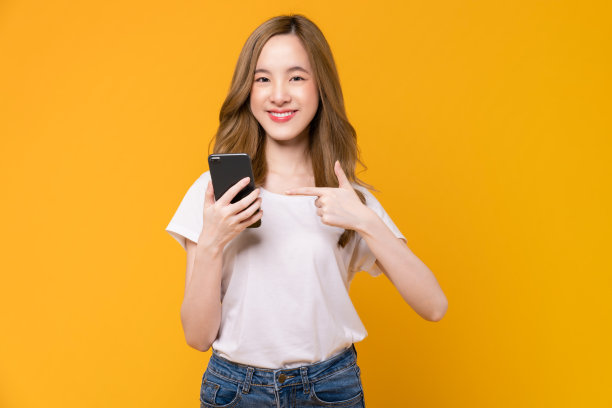 美丽的亚洲女人拿着智能手机，用手指指着黄色背景的手机。图片下载