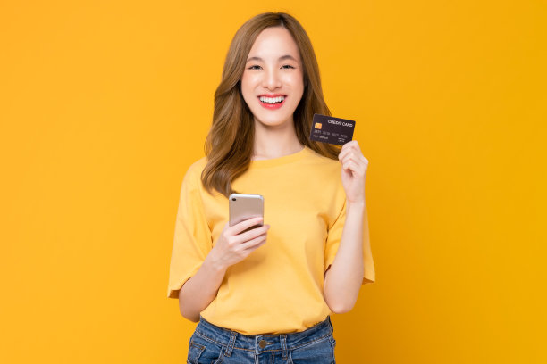 开朗美丽的亚洲女人拿着智能手机和橙色背景的信用卡模型。图片下载