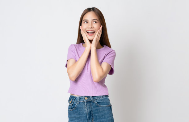 快乐微笑的年轻女子穿着随意的紫色t恤，手摸着脸，孤立在白色的背景上。图片下载