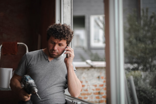 一个拿着智能手机和电钻的年轻人站在窗前。图片下载