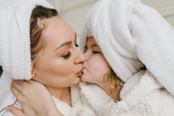 快乐的妈妈拥抱和亲吻孩子。快乐的女孩在家里度过美丽的一天。年轻的母亲带着小女儿穿着浴袍，头上披着毛巾，可爱地坐在床上。保健和家庭概念。特写镜头。图片下载