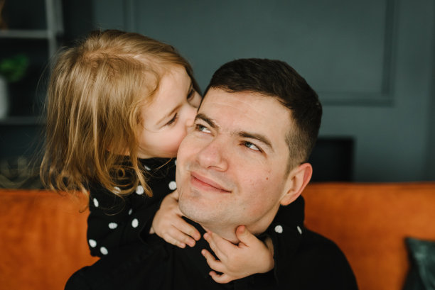 女儿亲吻并祝贺父亲(特写)。孩子微笑着拥抱爸爸，放松地躺在家里客厅的沙发上。爸爸和孩子笑着拥抱着。祝您父亲节快乐。家庭度假。图片下载