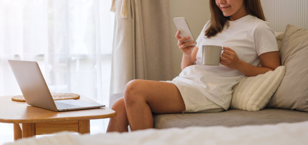一个年轻女子在家里用笔记本电脑一边喝咖啡，一边用手机聊天的特写图片下载