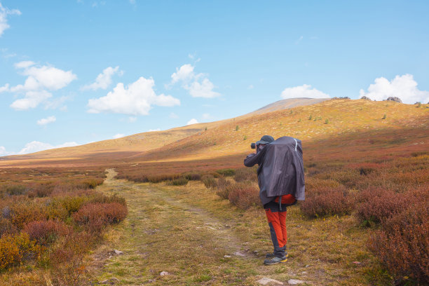 一个人穿着雨衣，背着大背包，在高山高原凋零的秋天植被中拍摄自然。背包客带着相机在多变的天气里拍摄山景图片下载