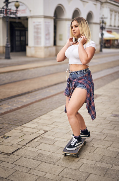 快乐的少女在城市街道上玩滑板，同时听着耳机上的音乐-保持微笑，享受生活的概念-背光夏日生活方式照片图片下载