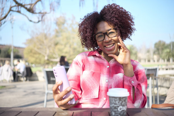 一个留着非洲式发型、戴着眼镜的黑人女孩在酒吧的露台上喝咖啡，对着镜头微笑的照片。图片下载