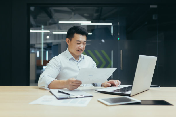 办公室工作。一位年轻的亚洲男子坐在桌子旁，正在用笔记本电脑和文件工作图片下载