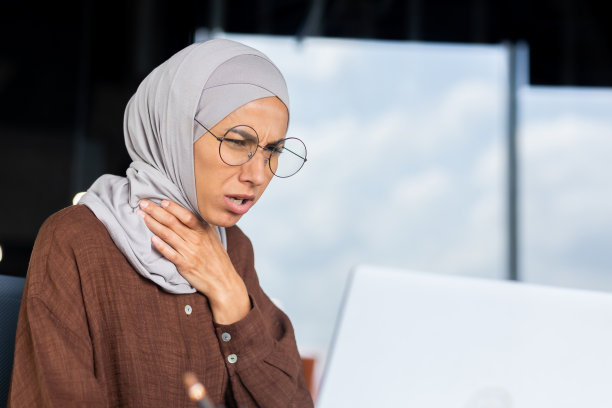 特写照片：戴着头巾的年轻阿拉伯女人在办公室用笔记本电脑工作，手扶着脖子，感到喉咙剧痛、嗓子疼、发烧、咳嗽图片下载