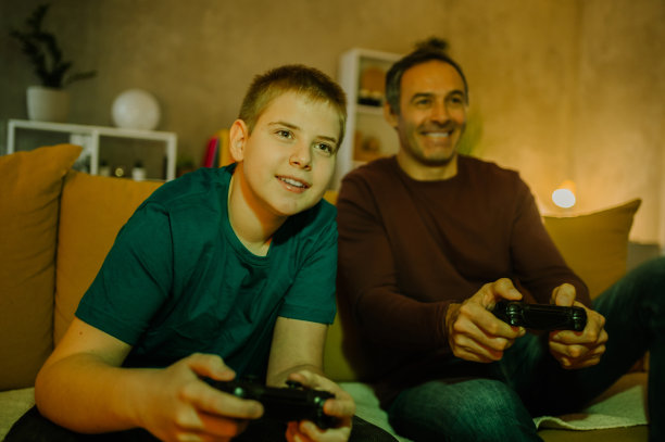 一个金发少年和他父亲一起玩电子游戏，享受他的空闲时间。图片下载
