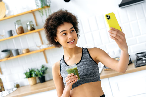 健康强化绿色奶昔。健康饮食，素食主义，生食。非裔美国女孩，博主，用智能手机，拿着一杯有机奶昔，微笑，自拍或录制视频流图片下载