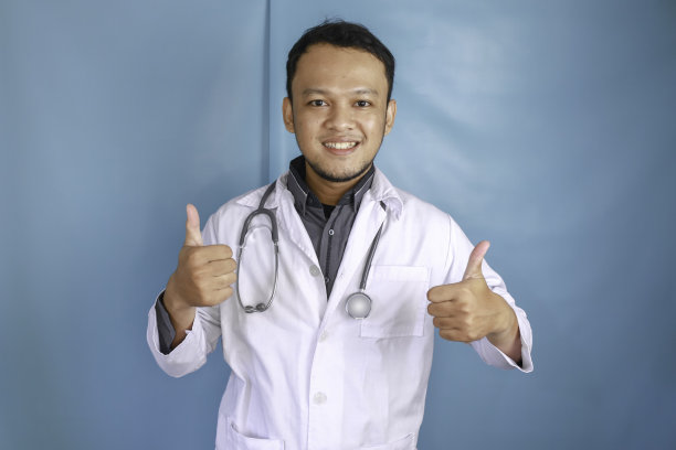 一个年轻的亚洲男性医生的肖像，一个医疗专业人员微笑着，在蓝色的背景上显示大拇指或OK的标志图片下载