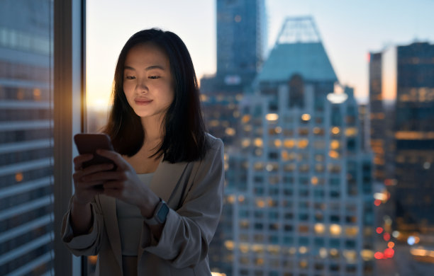 年轻的亚洲女商人站在晚上的城市办公室窗口使用电话。图片下载