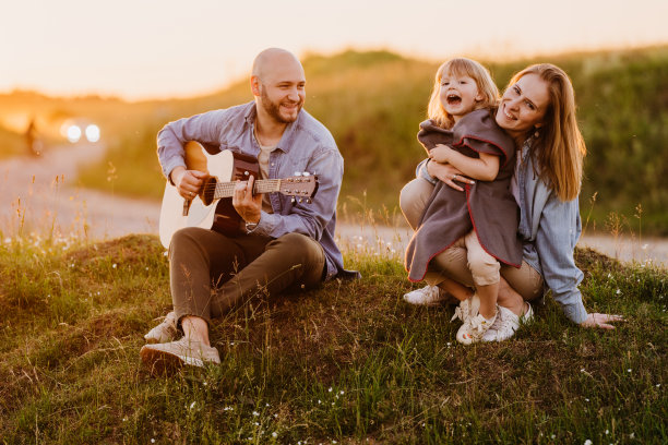 一家人在大自然中享受日落时光。大胡子的年轻成年男子穿着休闲装，坐在农场的小山上为妻子和女儿弹吉他。欢快的金发妈妈拥抱着正在唱歌的小女孩。图片下载