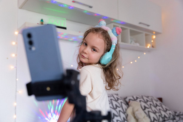 一个戴着耳机的小女孩在房间里用手机录制视频或故事，背景是彩灯和手电筒。内容创建者记录vlog。图片下载
