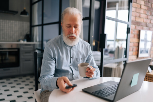 在现代厨房里，大胡子的成熟老人在笔记本电脑和智能手机上工作，拿着一杯咖啡，坐在桌子旁图片下载