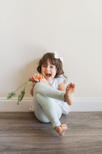 一个快乐的4岁古巴裔美国女孩，棕色卷发，棕色眼睛和橄榄色皮肤，穿着白色衬衫，上面有兔子的青色打底裤和白色头发蝴蝶结，赤脚在家里吃一整根胡萝卜，在一个中性的简单空间里图片下载