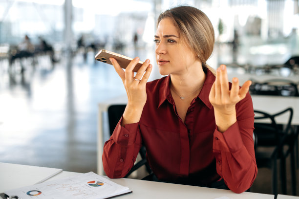 专注、忙碌、成功、自信的白人商务女性，坐在办公室的办公桌前，用她的智能手机，给客户录制语音信息，用免提与同事通话，然后看向别处图片下载