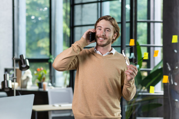 一个年轻的自由职业者正站在办公室里讲电话。他手里拿着眼镜，微笑着看着镜头图片下载