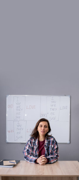 快乐的女老师坐在木桌前，看着背景是英语语法规则的白板前的镜头。女教师讲师录制教育视频，教导学生图片下载