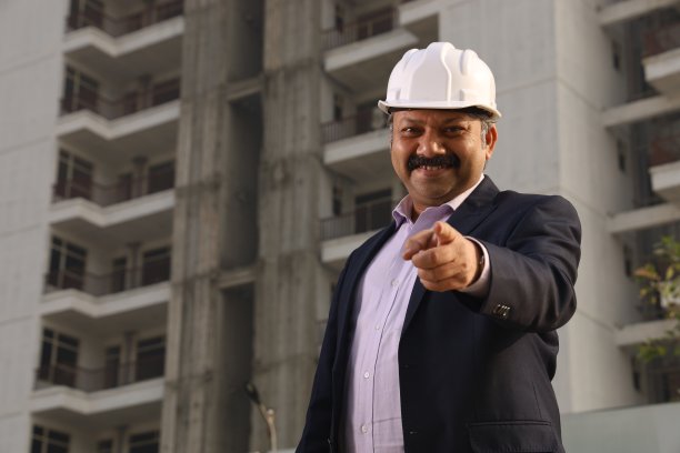 一个快乐的印度建筑工人戴着建筑工人的头盔站在一个建筑商的财产前。图片下载