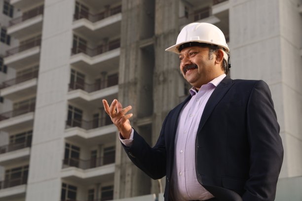 一个快乐的印度建筑工人戴着建筑工人的头盔站在一个建筑商的财产前。图片下载