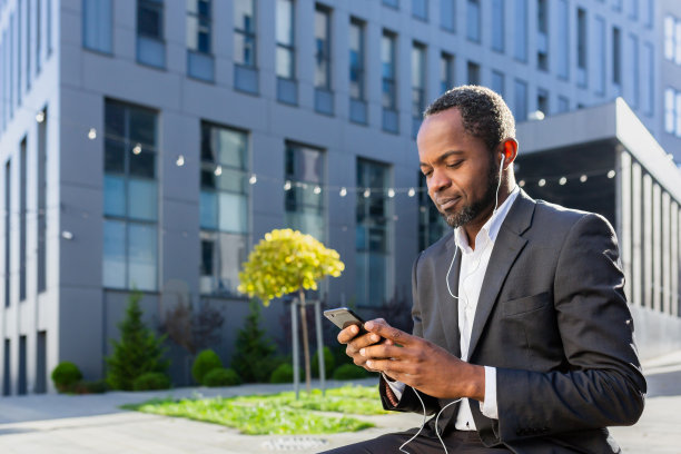 一个资深的非裔美国人，一个穿着西装的商人，坐在办公中心外面，戴着耳机，在手机上听音乐、播客和有声读物图片下载