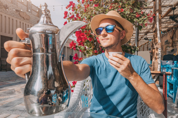 在迪拜一家熙熙攘攘的咖啡馆里，一名男子沉浸在该地区的文化和历史中，他从传统的咖啡壶里倒了一杯芳香的阿拉伯咖啡，细细品味着每一口。图片下载