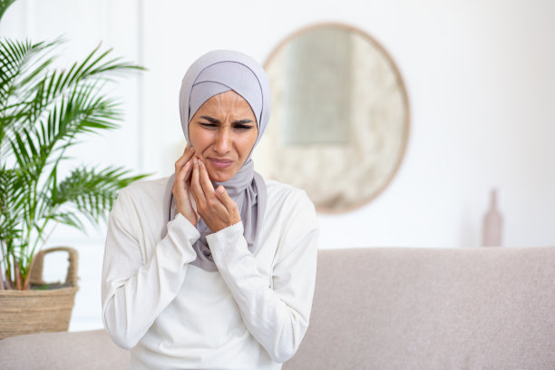 戴着头巾的穆斯林年轻女子在家中、酒店里坐在沙发上，手托着脸颊，感到严重的牙痛、不适，需要牙医的帮助图片下载