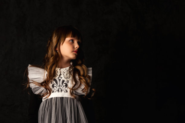 肖像封面上的小女孩在人偶形象中被孤立在阴影中，眼神呆呆地望向远方。迷人的孩子9-10岁卷曲的头发在黑色背景。时尚的风格理念。复制广告文本空间图片下载