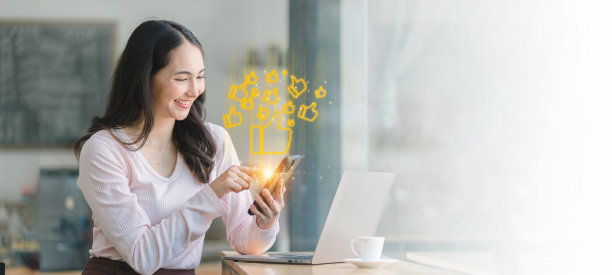 漂亮的亚洲女商人坐在咖啡馆里，用智能手机，对着抽象的灯光和竖起大拇指的社交图标微笑。图片下载