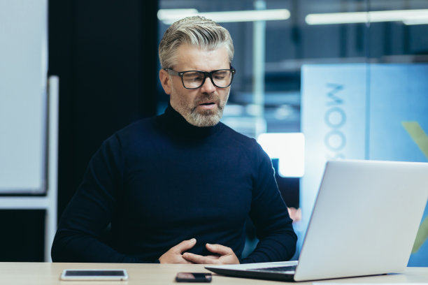 生病的白发商人中毒，老板抱着肚子，有严重的胃痛，在现代办公室工作的成熟男子使用笔记本电脑图片下载