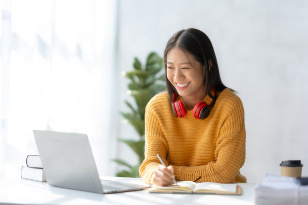 亚洲女商人坐在家里做笔记，用笔记本电脑愉快地工作。年轻的亚洲女学生在图书馆做笔记和使用笔记本电脑。图片下载